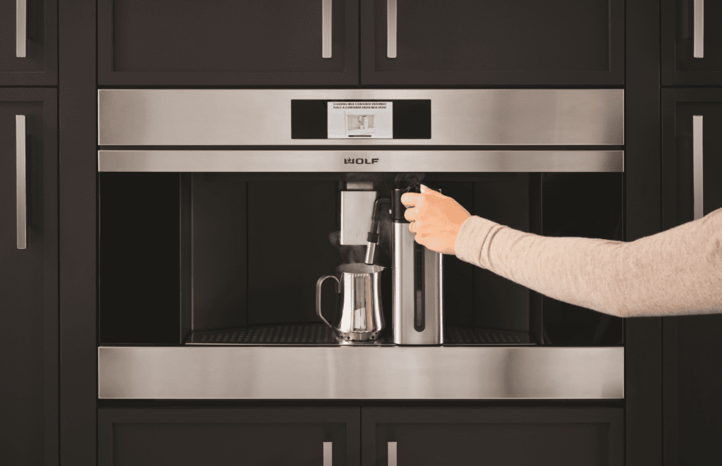 Close-up of a modern Wolf coffee system built into black cabinetry, with a hand preparing a beverage.